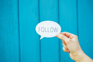 hand holding small "follow" sign in quotes against blue fence backdrop