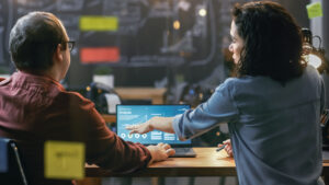 Male and Female Financiers Look at Statistics and Graphs Shown on the Laptop, They Talk Solving Problems. In the Background Creative Office.